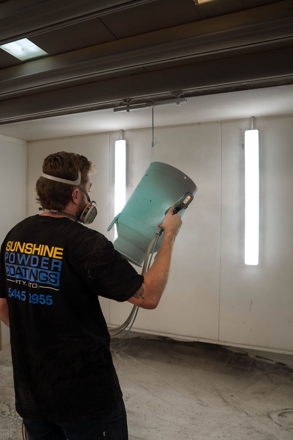 A person wearing a respirator sprays a light blue metal object inside a professional powder coating booth.