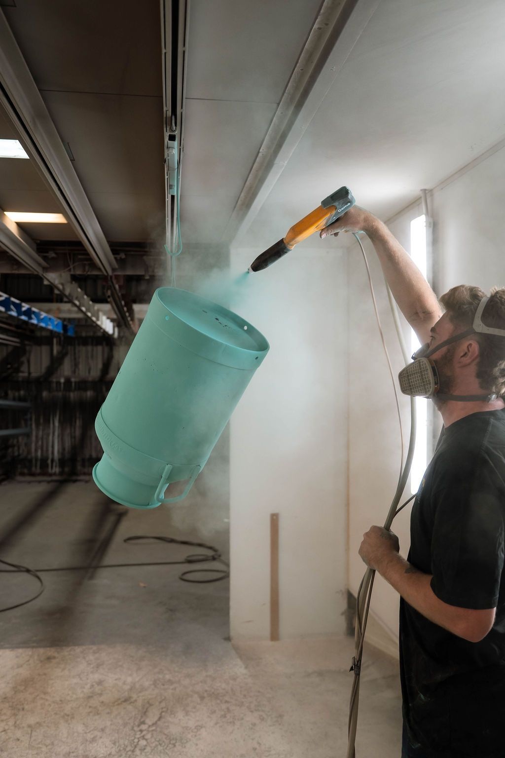 A person wearing a respirator sprays turquoise powder coating onto a metal container in an industrial workshop.