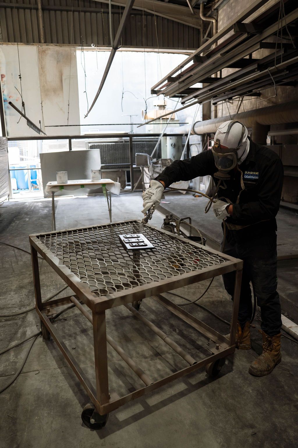 A worker in a respirator and protective gear sands a small metal plate on a workshop table.