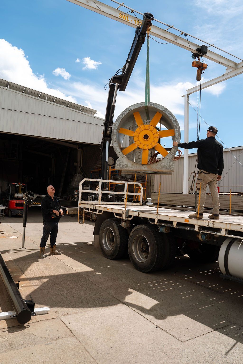 A crane lifts a large industrial fan on a truck bed, while two people stand nearby on an outdoor industrial lot.