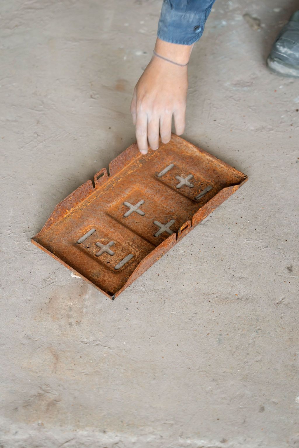 A person's hand reaches toward a rectangular, rusted metal tray featuring embossed plus signs on a concrete floor.