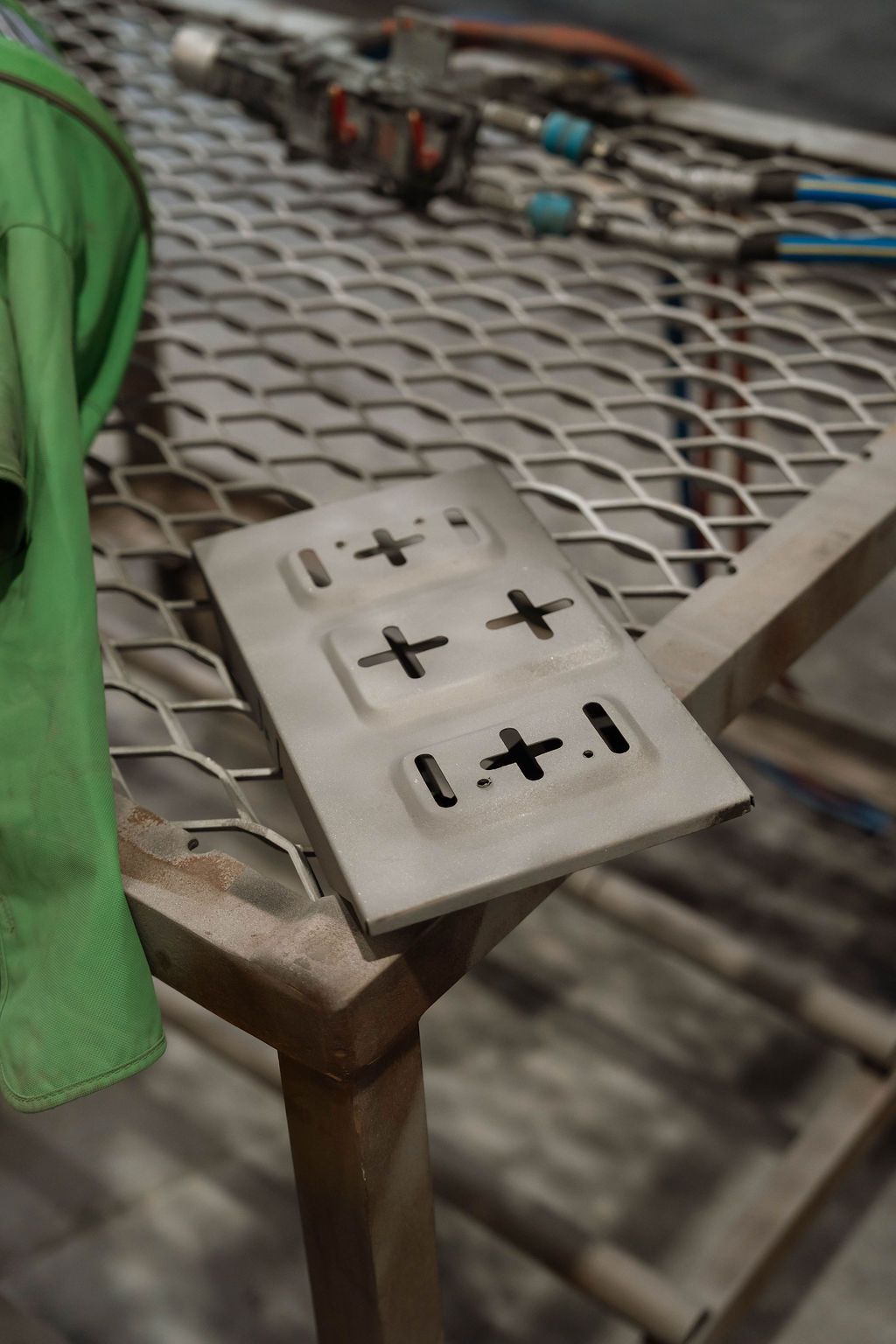 A metal plate with three cross-shaped cutouts, resting on a metal mesh industrial workbench next to a green fabric.