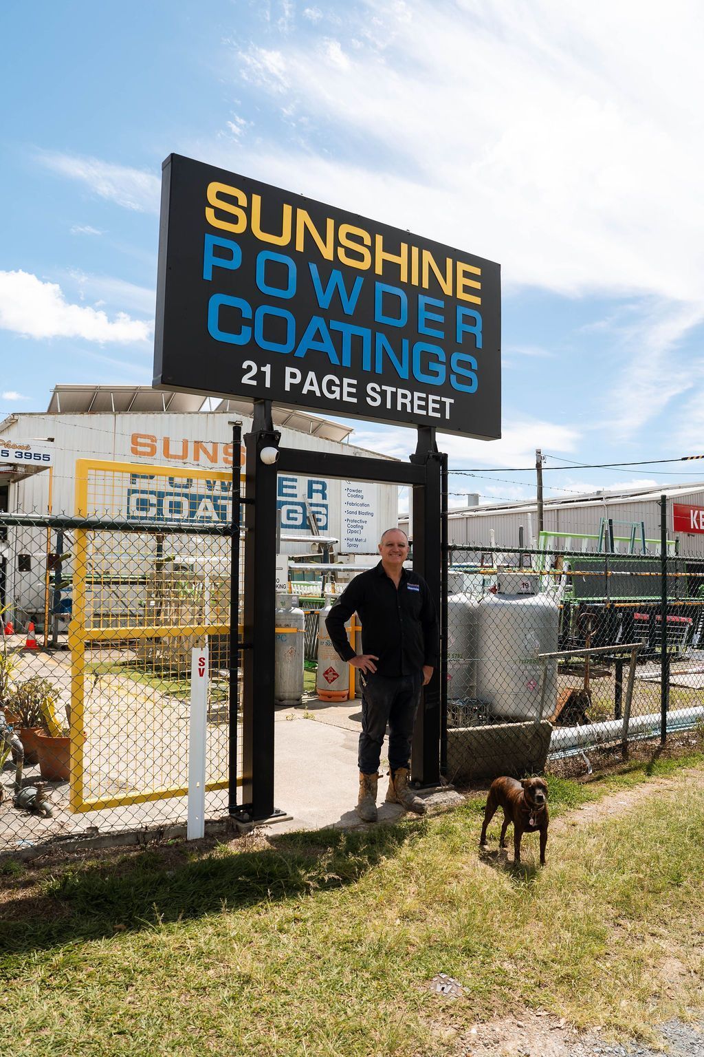 A person stands near a dog in front of a Sunshine Powder Coatings sign at 21 Page Street on a sunny day.