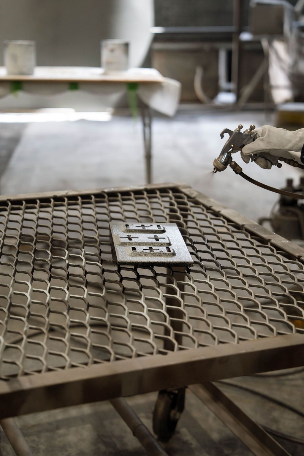 A person wearing a glove spray-paints a small metal component on a metal grate workbench in a workshop.