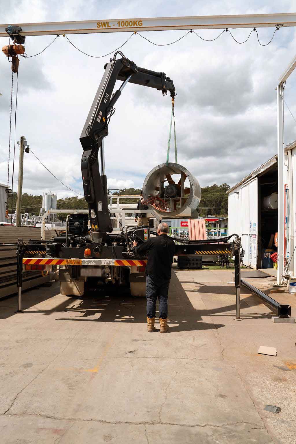Crane truck lifting a large metal ring in an industrial yard with a worker standing nearby