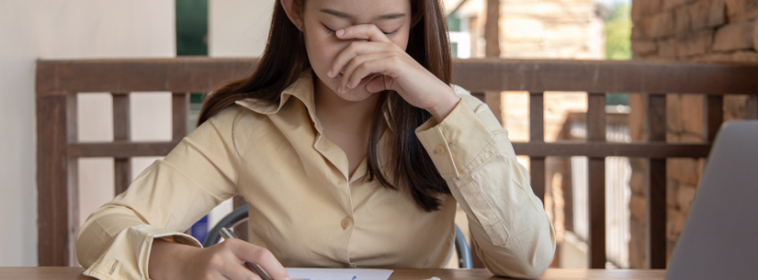 Woman sitting at a table, rubbing her nose with one hand while writing in a notebook, laptop nearby