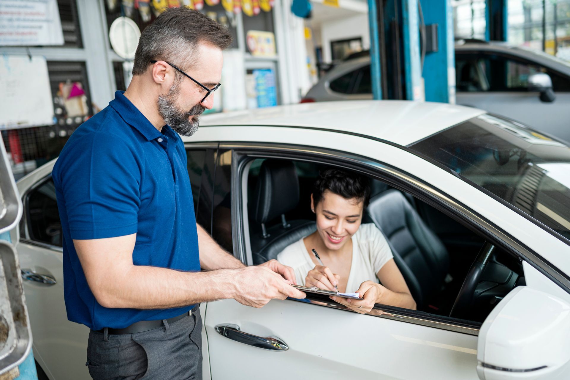Mechanic handing paperwork to a driver in a white car, in an auto repair shop.