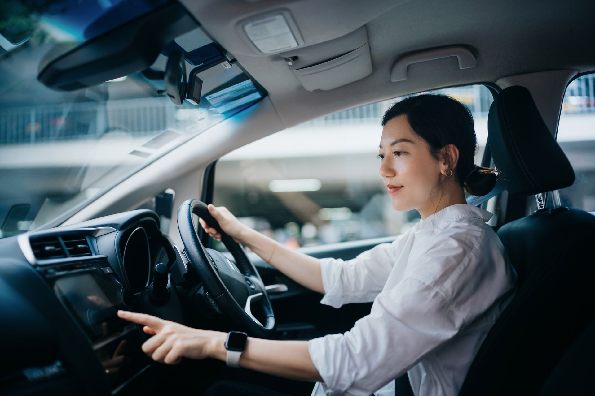 Woman in white shirt inside car, touching touchscreen while driving.