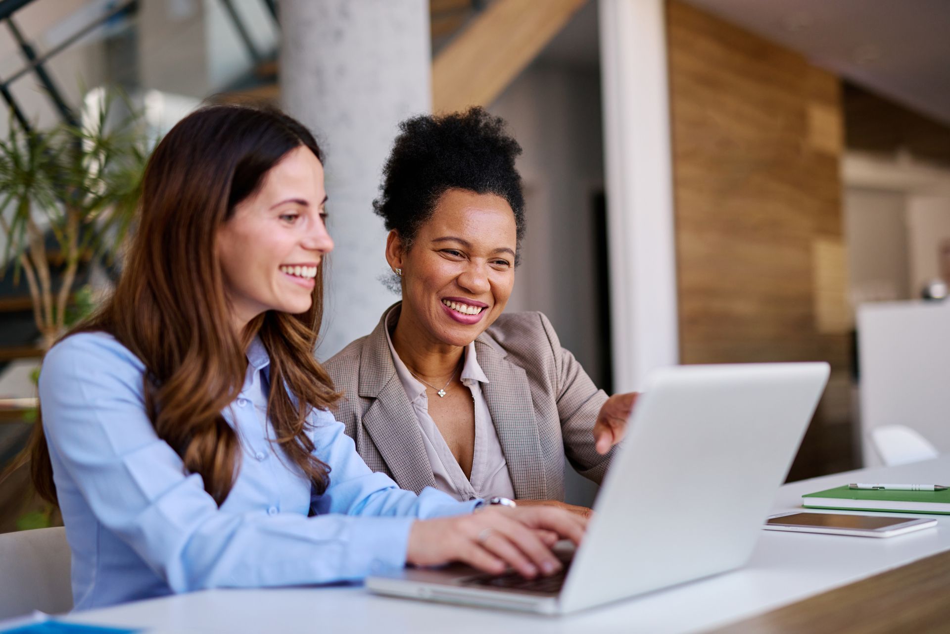 Two women smiling while looking at a laptop in a modern office. One points at the screen.