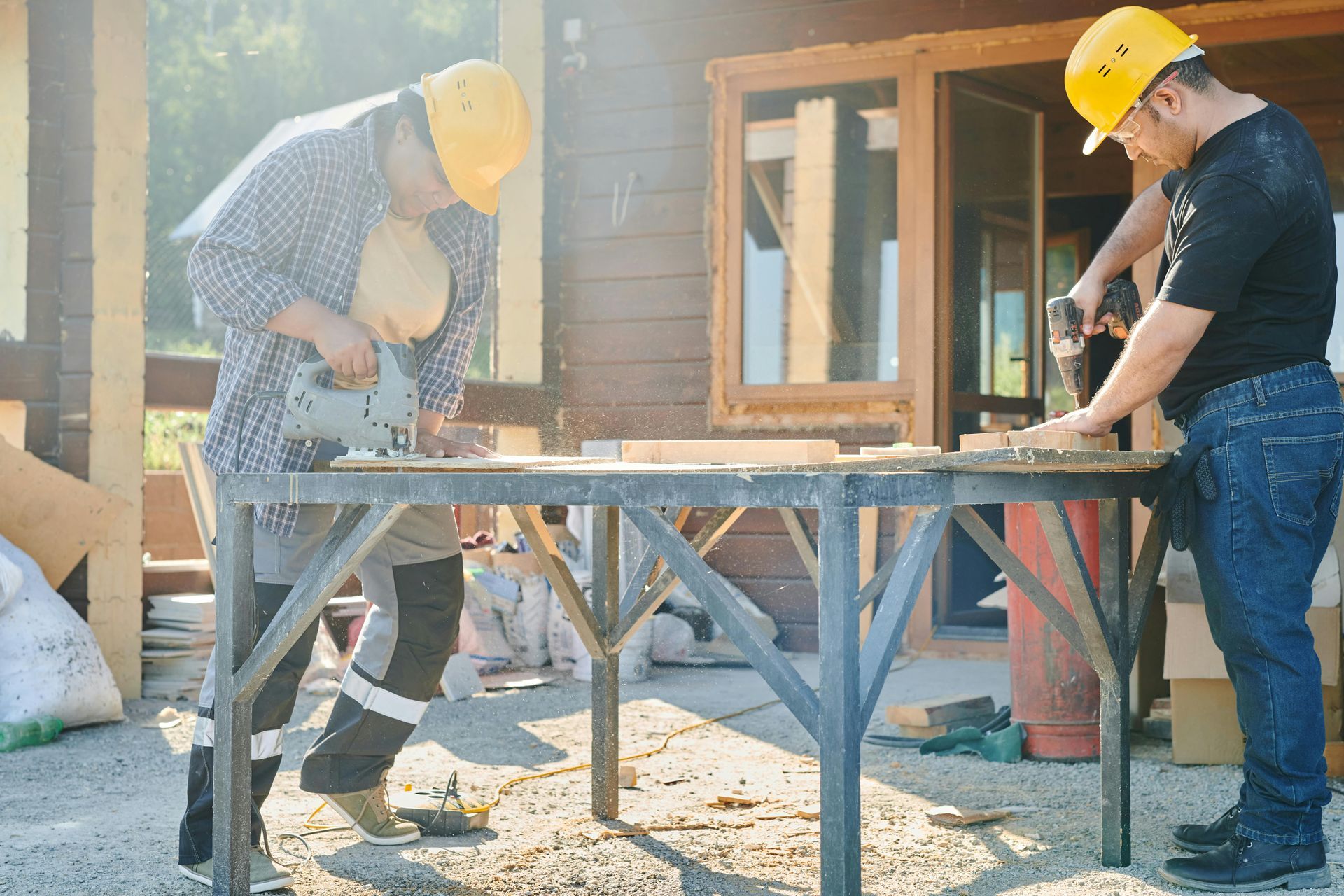 Two construction workers wearing hard hats cutting wood outdoors.