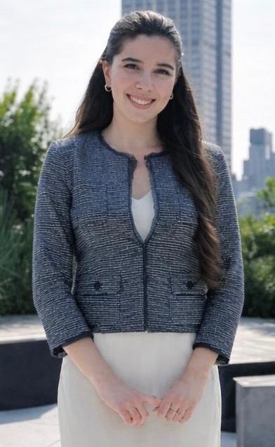 Woman standing outdoors in a gray jacket and white dress, smiling with city buildings in the background.