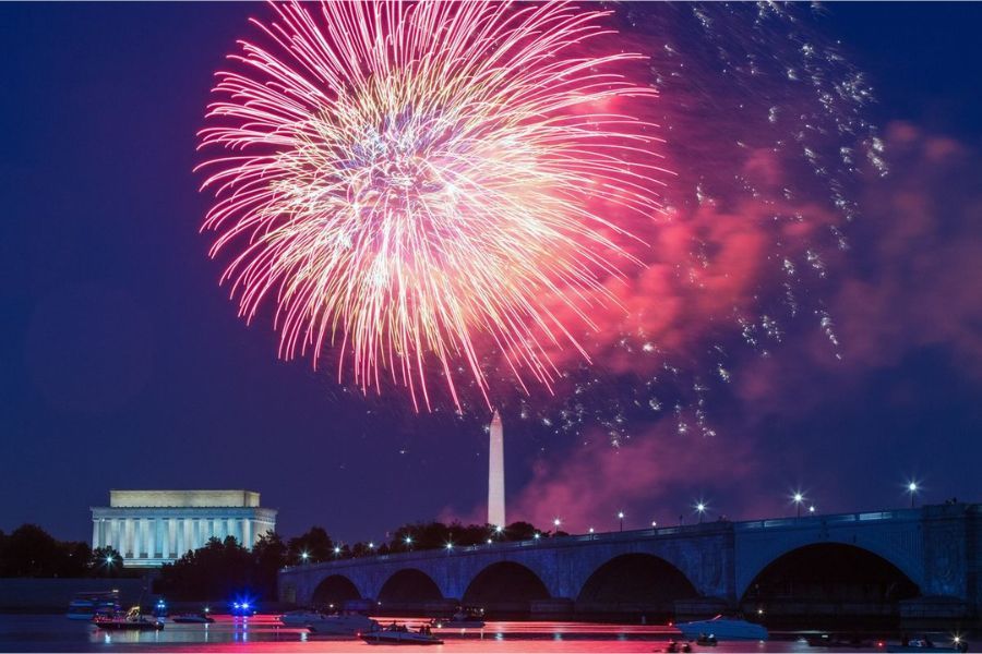 Fireworks burst over a bridge and monument at night, with pink and white light filling the sky.