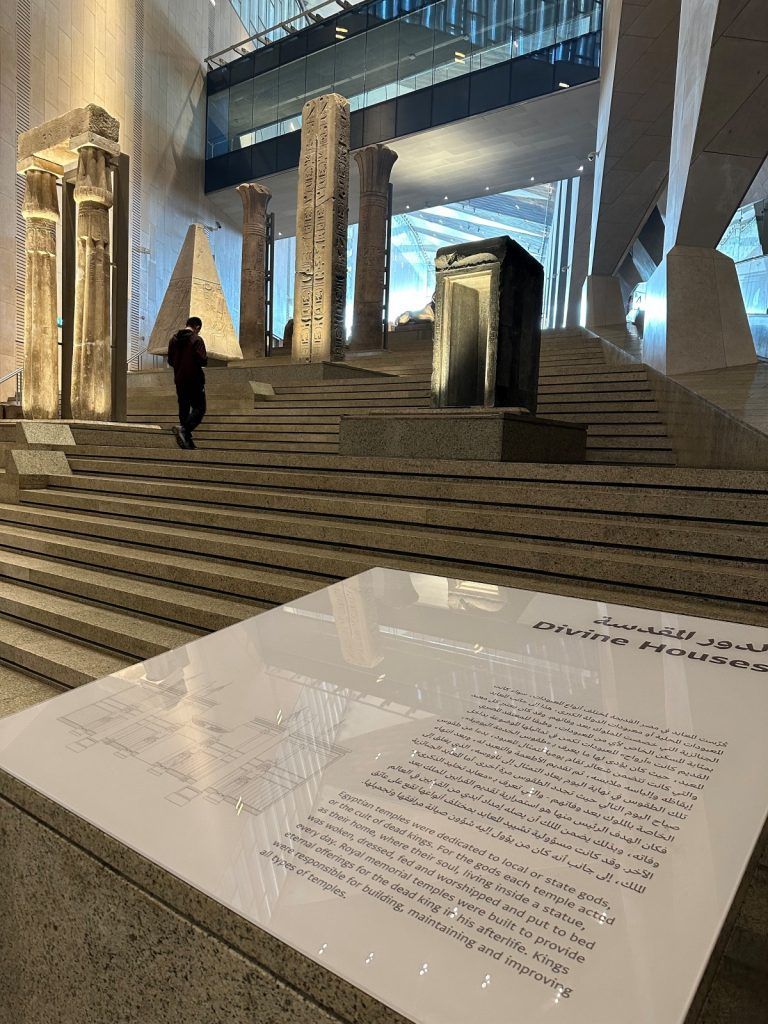Museum exhibit with ancient columns and artifacts; a person walks up stairs, modern architecture backdrop.