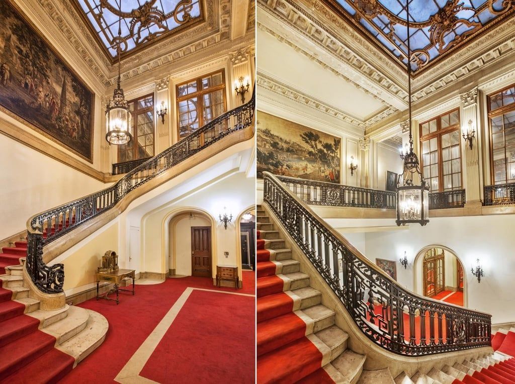 Ornate staircase with red carpet and dark railing in a grand building. Tapestries and detailed ceiling.