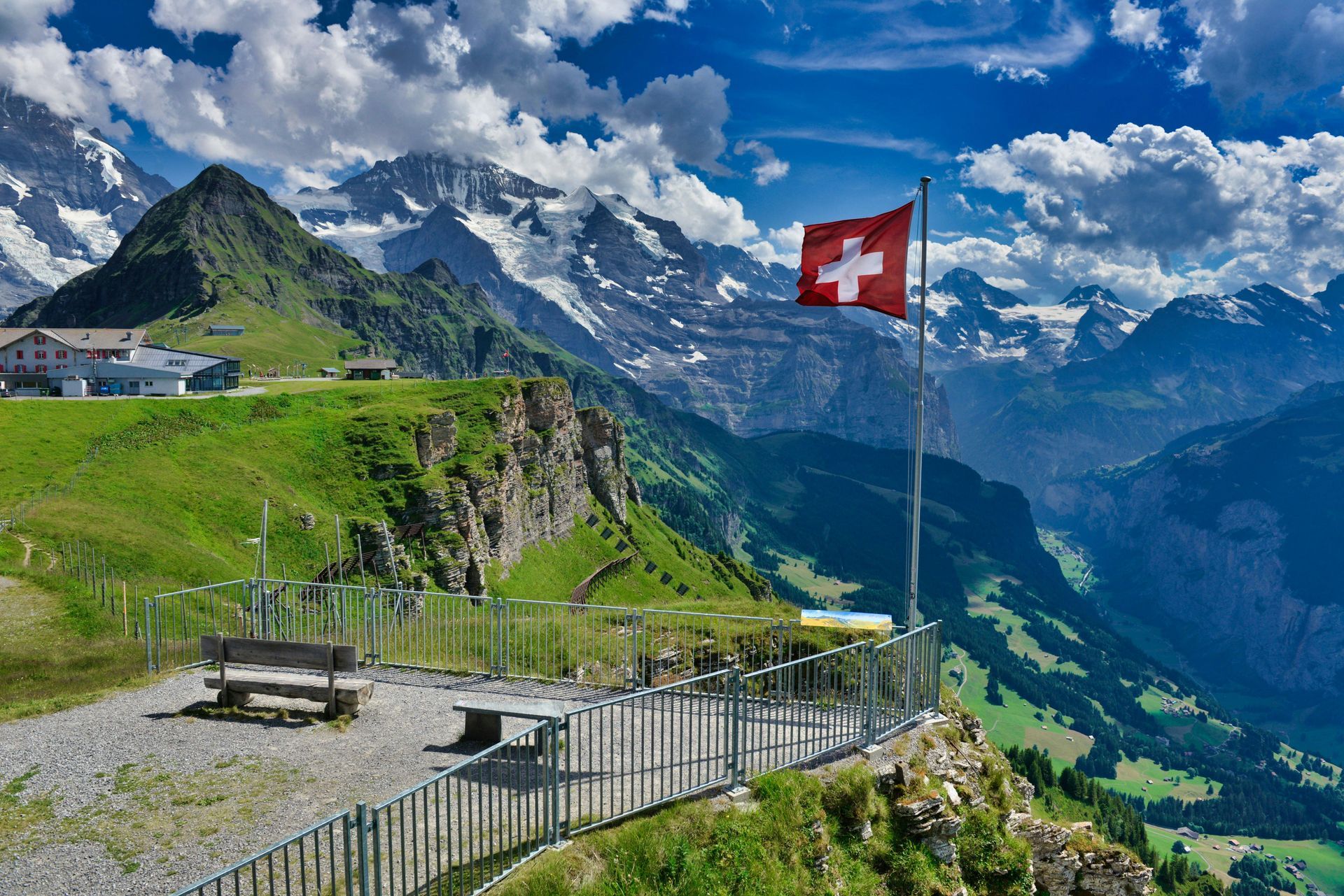 Swiss flag on a mountain overlook with benches, green hills, and snowcapped Alps under a blue sky