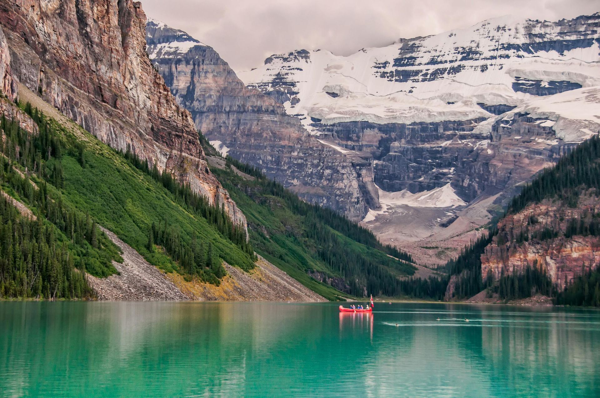 Turquoise mountain lake with snow-capped peaks, forested slopes, and a small red canoe