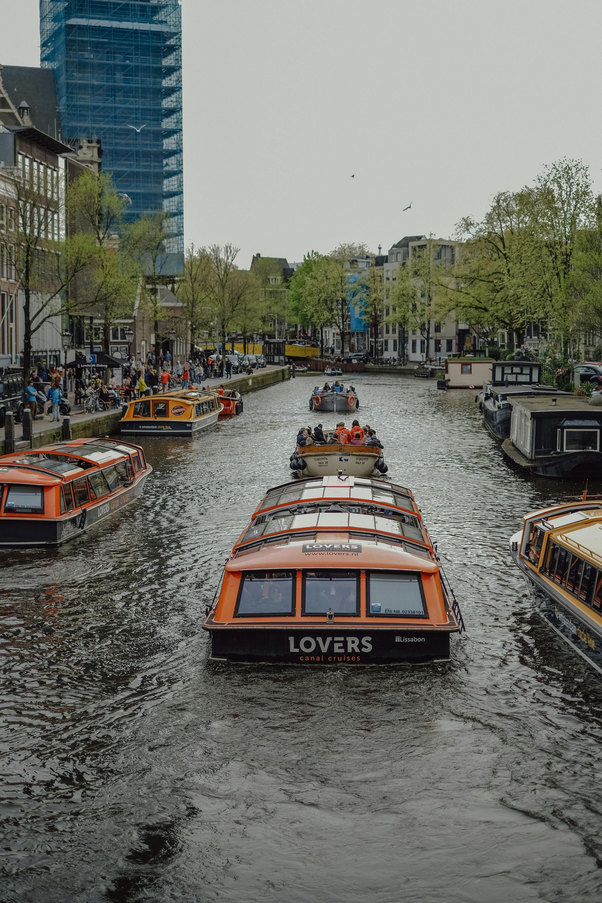 Canal boats lined up on a city canal, with trees, buildings, and spectators along the banks.