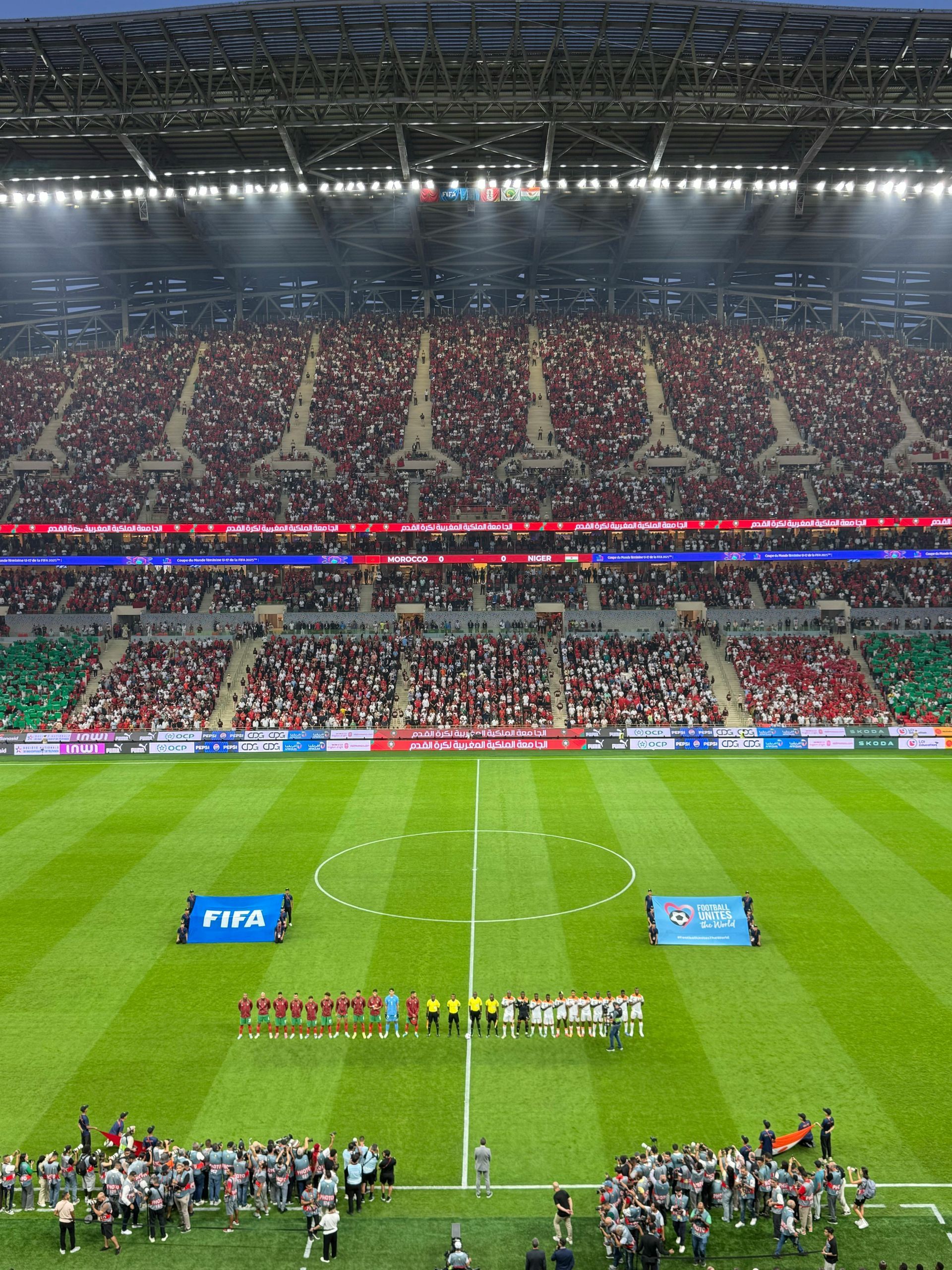 Soccer stadium packed with fans, teams lined up at midfield, and a bright green pitch under stadium lights.