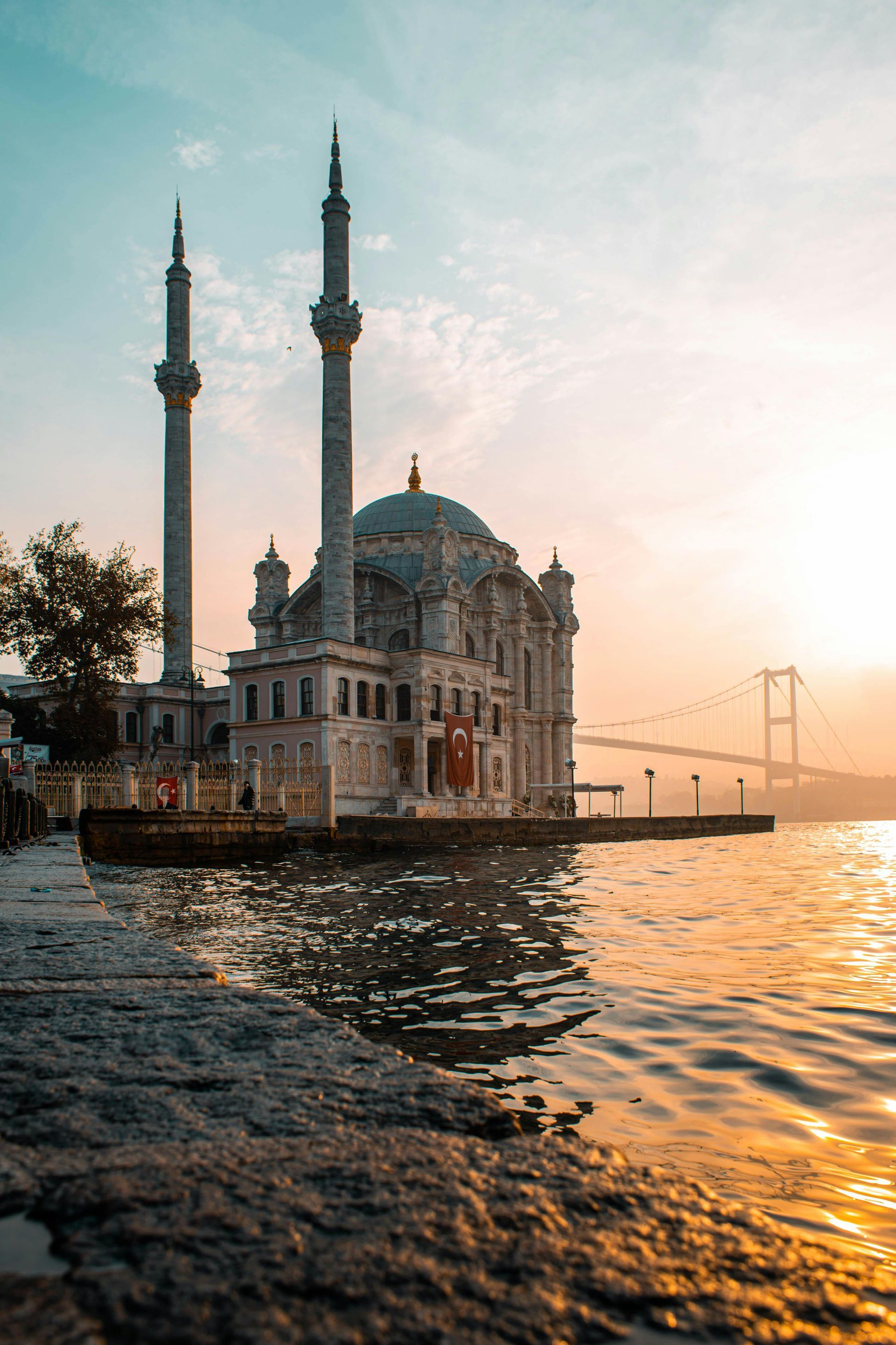 Mosque at sunset beside rippling water, with warm golden reflections and a bridge in the background