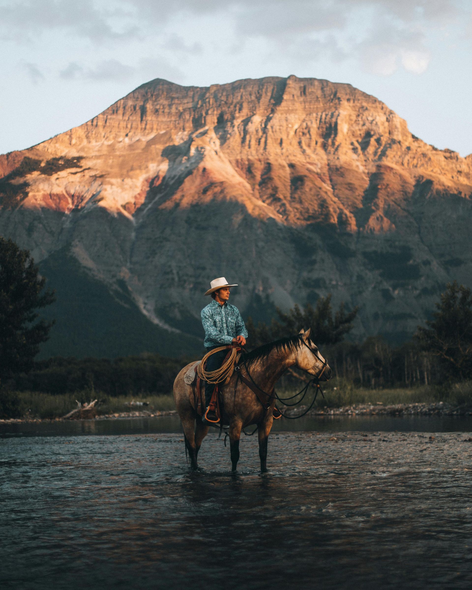 Rider on horseback crossing a shallow river at sunset with a mountain lit by warm light in the background