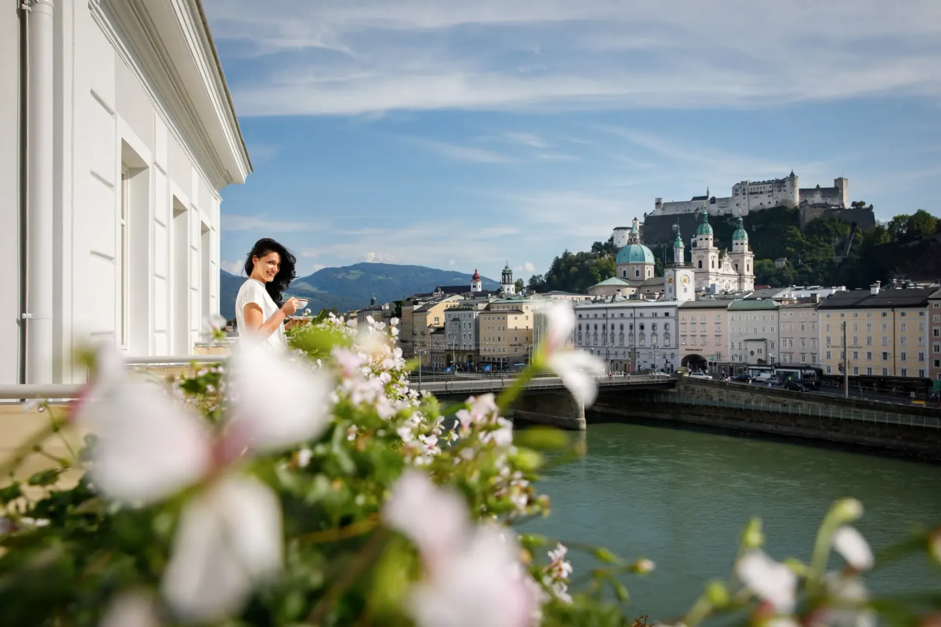 Riverfront cityscape with flowers in the foreground and a person leaning by a white building overlooking a castle on the hill