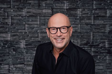 Lächelnder, glatzköpfiger Mann mit Brille und schwarzem Hemd vor einer dunklen Steinmauer