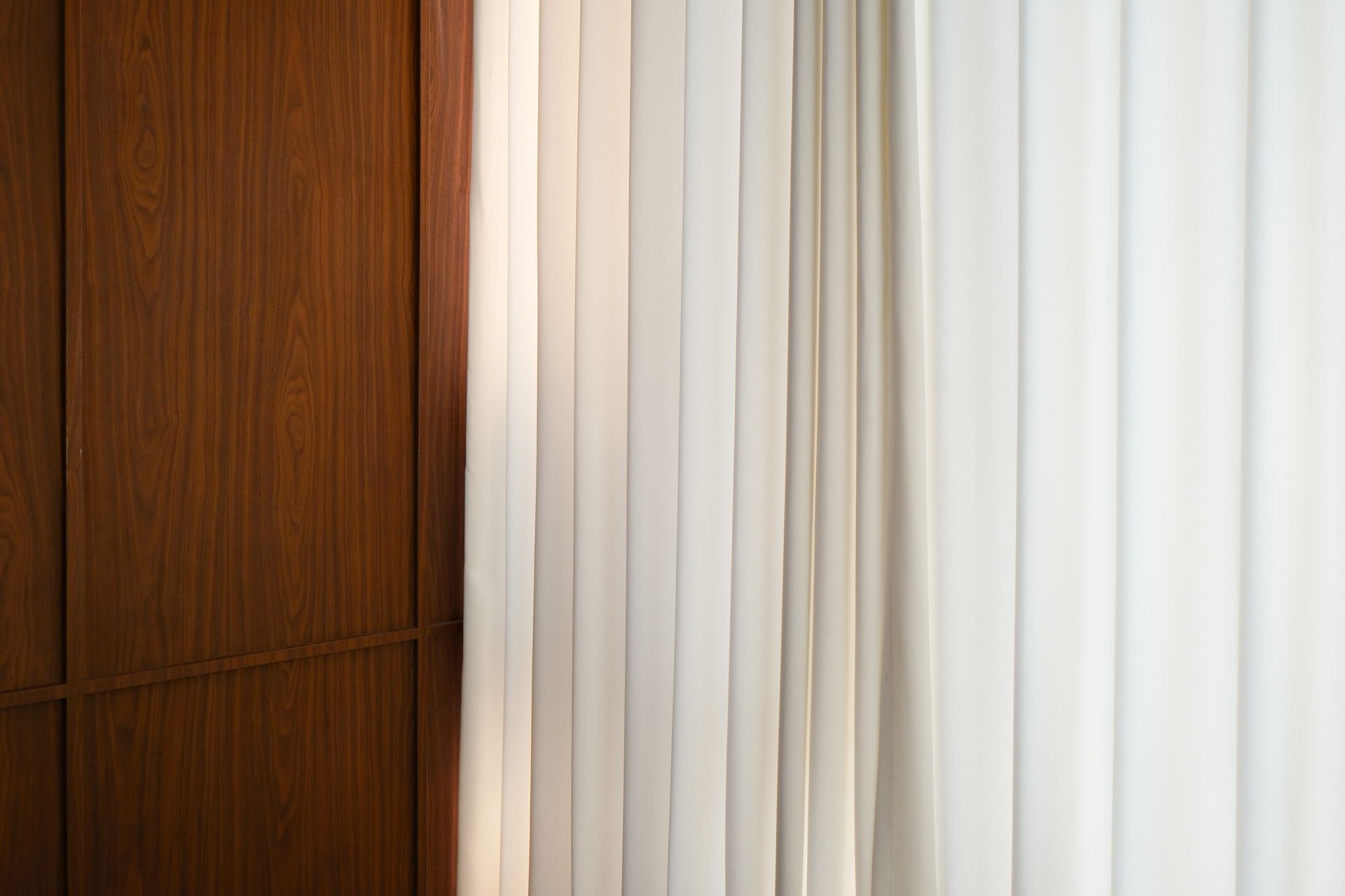 Vertical white fabric blinds hanging beside a wooden wall or panel in an interior space.
