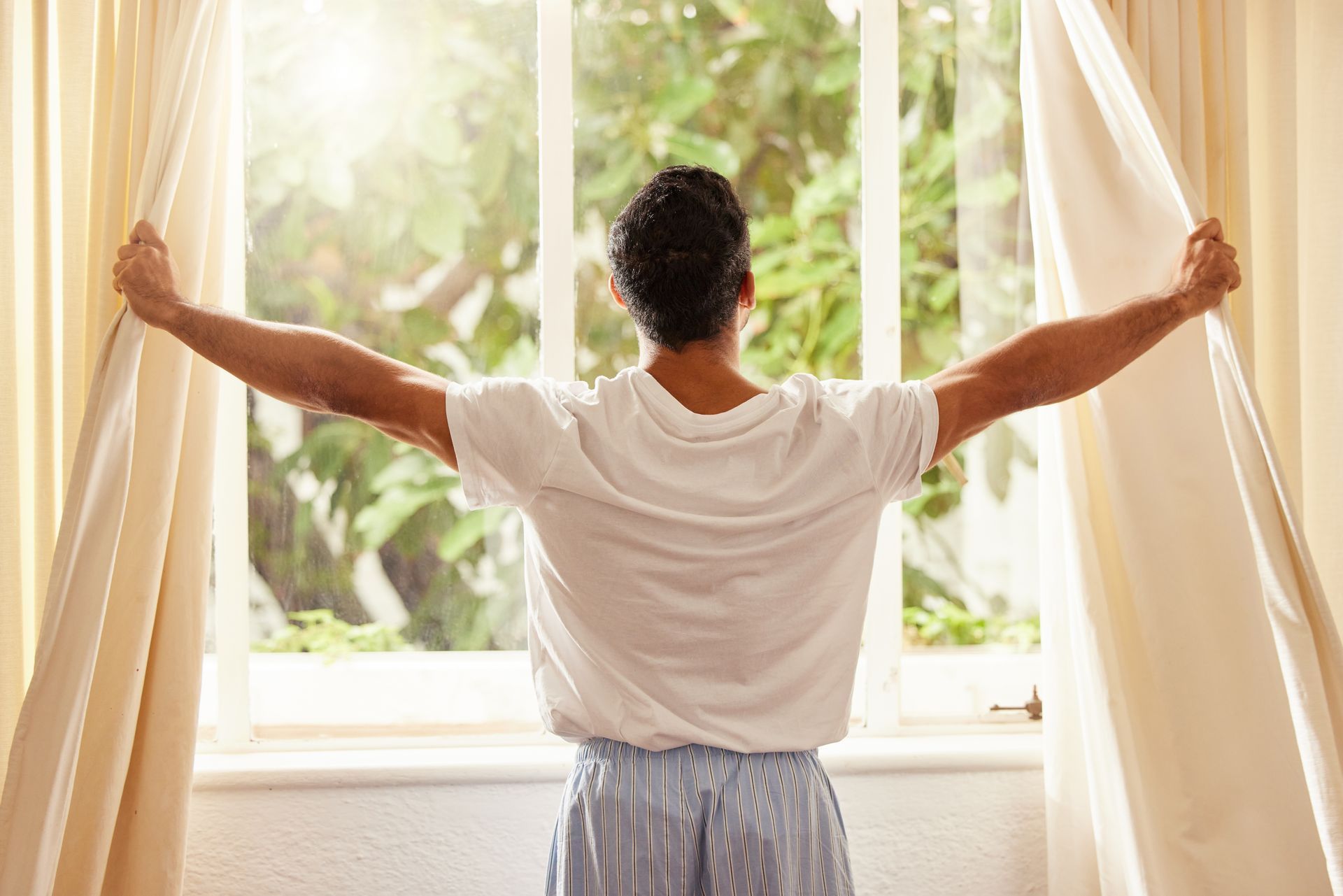 A person opens drapes; sunlight streams in, filling the room with warmth and serenity.