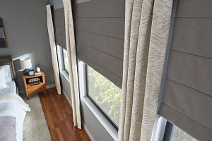 A bedroom with a bed , nightstand , curtains and roman shades on the windows.