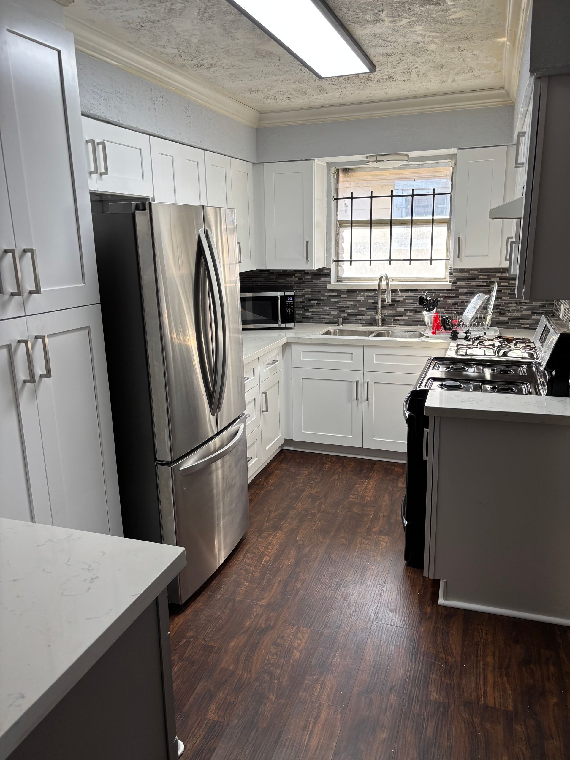 White kitchen with stainless steel refrigerator, white cabinets, dark wood floor, and window.