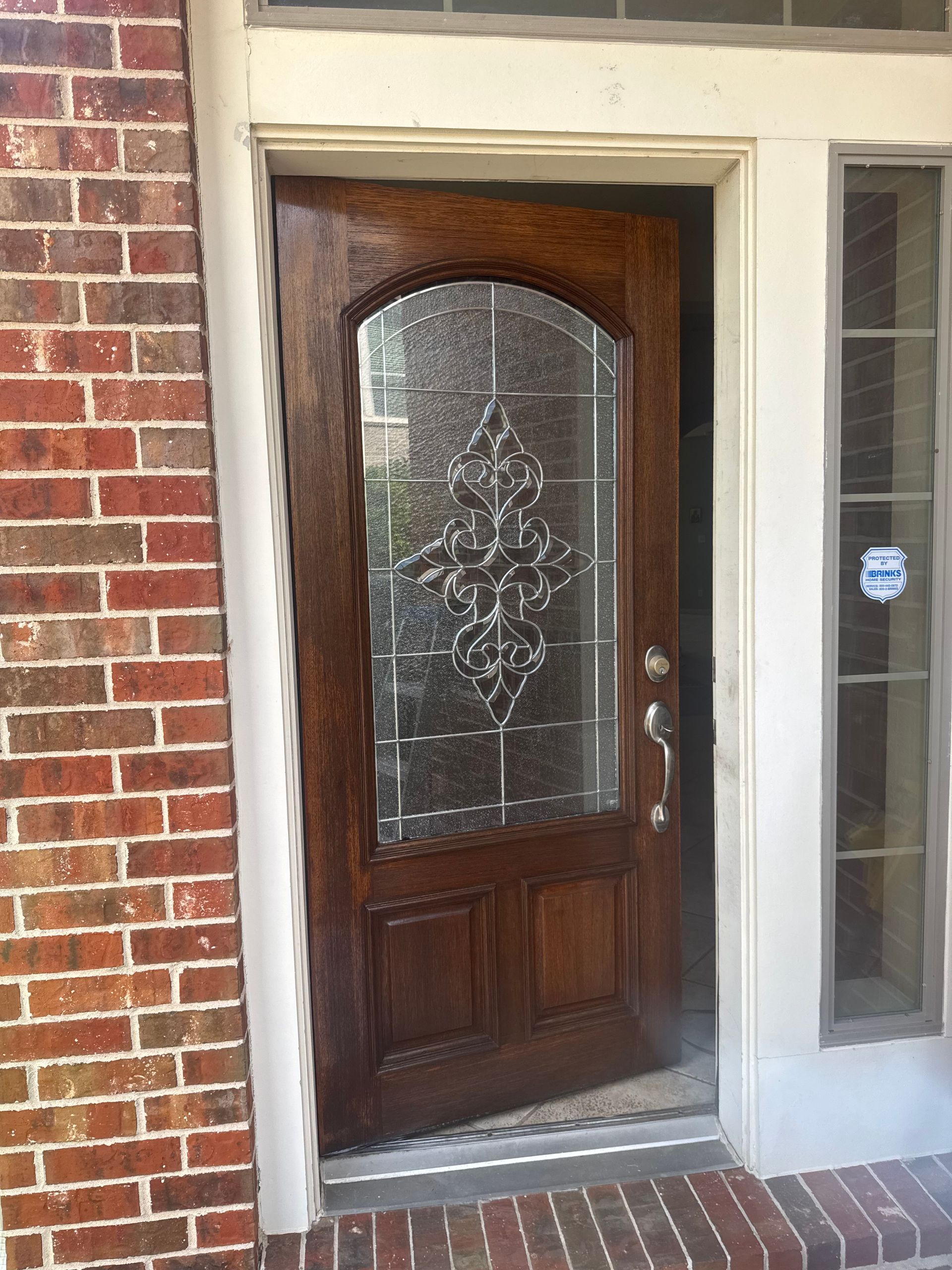 Wooden front door with decorative glass panels, partially open, surrounded by brick and white trim.