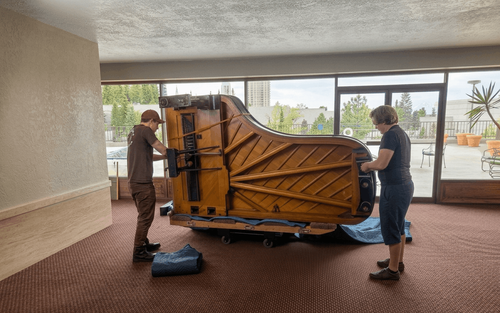 Two workers remove the legs of a large baby grand in an office space