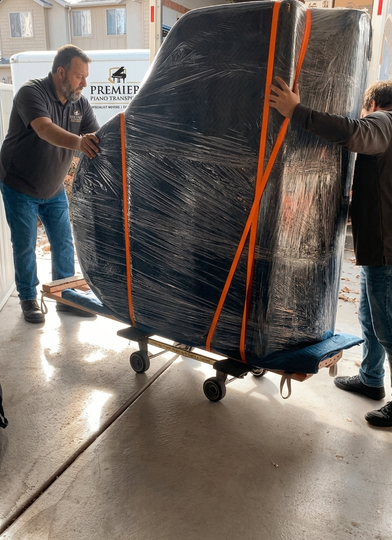 Two workers guide a wrapped baby grand on a moving dolley toward
