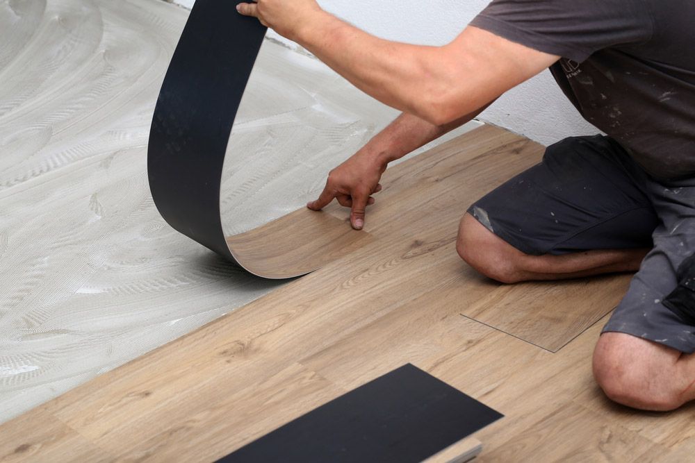 A Man is Kneeling Down While Installing a Vinyl Floor — Bayside Carpets & Flooring in Harrington, NSW