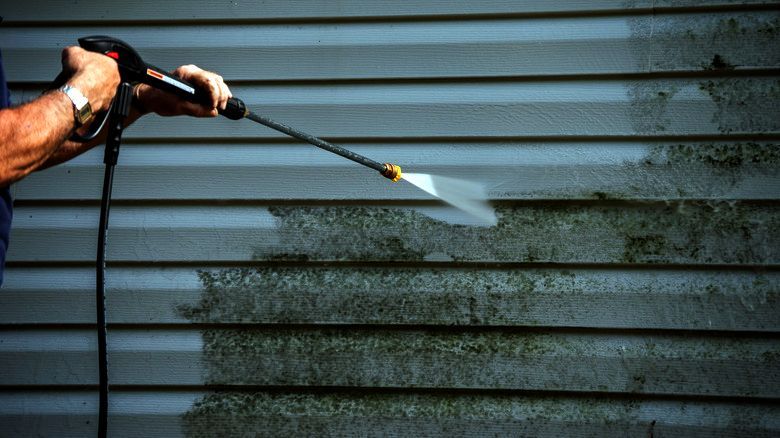 Person pressure-washing a gray house siding, spraying water onto a stained section.