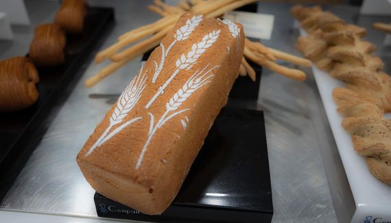 Una pagnotta marrone da dessert con glassa bianca a forma di foglia su un vassoio da esposizione, con i pasticcini sfocati sullo sfondo.
