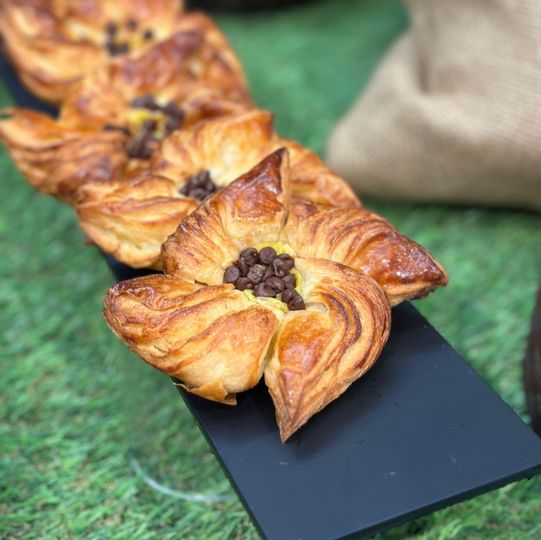 Pasticcini dorati a forma di fiore, ricoperti di gocce di cioccolato, disposti su un vassoio scuro sopra un telo verde.