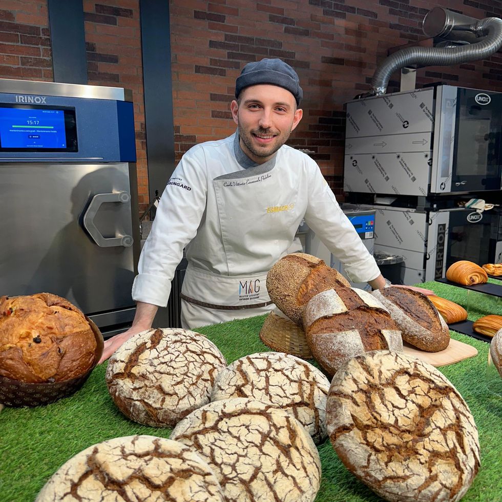 Un fornaio in cucina, con un assortimento di pagnotte rustiche su un tavolo, sorride alla telecamera.