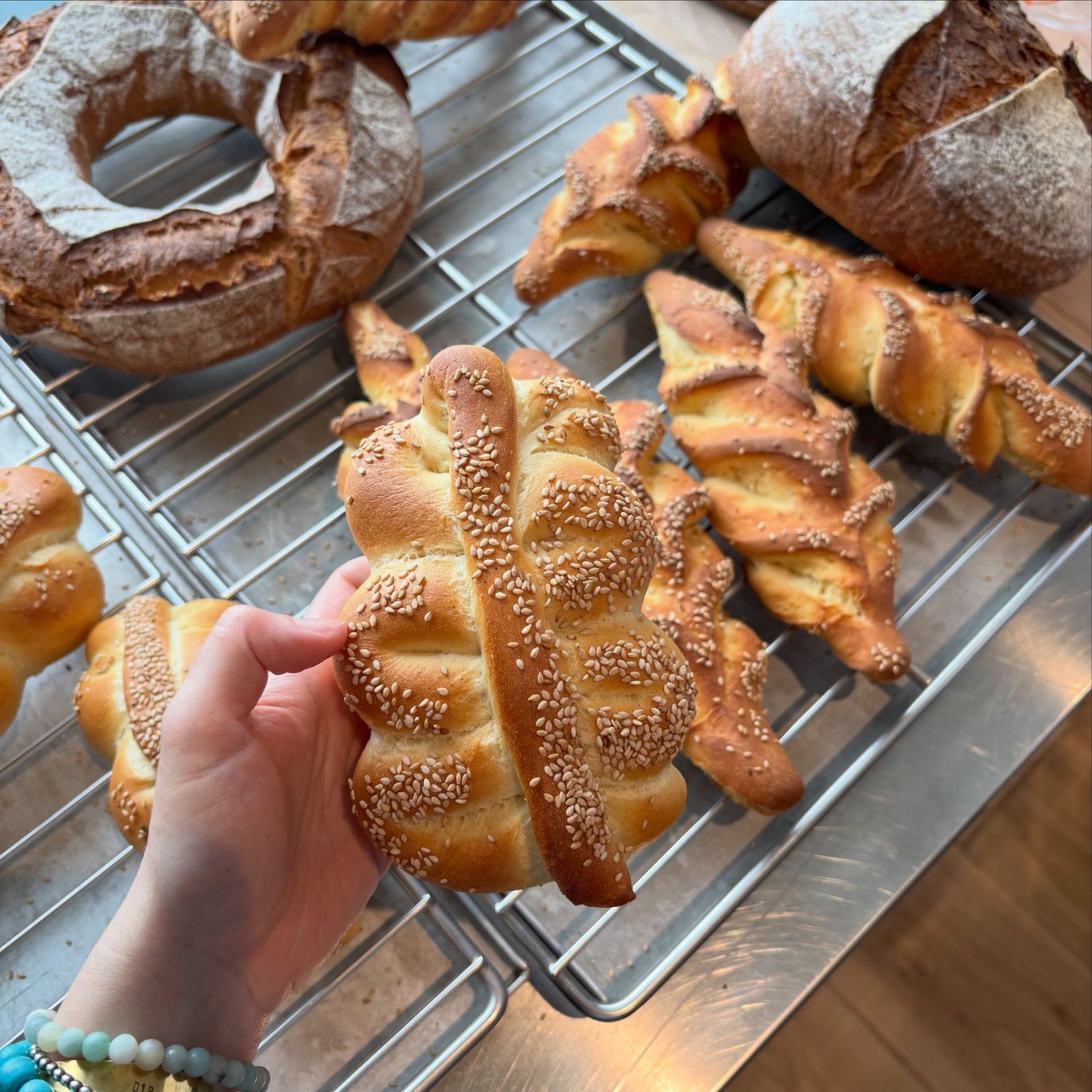 Una mano regge una treccia di pane al sesamo sopra una griglia di raffreddamento piena di pasticcini freschi assortiti.