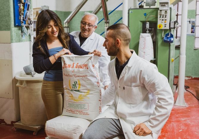 Un fornaio con il grembiule bianco tiene in mano una pagnotta rotonda davanti alla vetrina di un panificio.