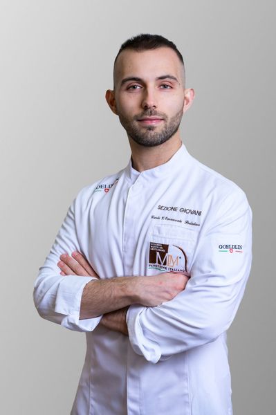 Chef in camice bianco e berretto nero sorridente mentre tiene in mano un panino in un ambiente di cucina.