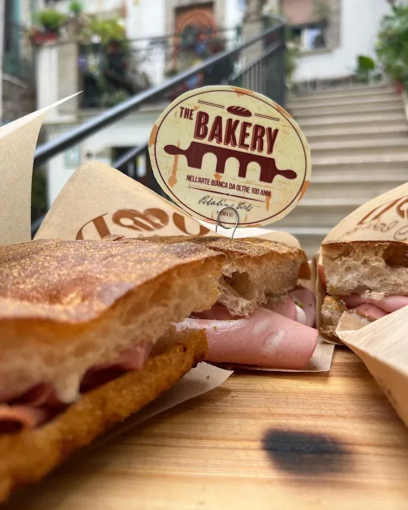 Panino da forno con prosciutto su tagliere di legno, insegna della panetteria sullo sfondo, davanti alle scale esterne