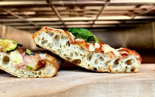 Una mano regge un panino intrecciato al sesamo sopra una griglia di raffreddamento con diversi tipi di pane fresco.