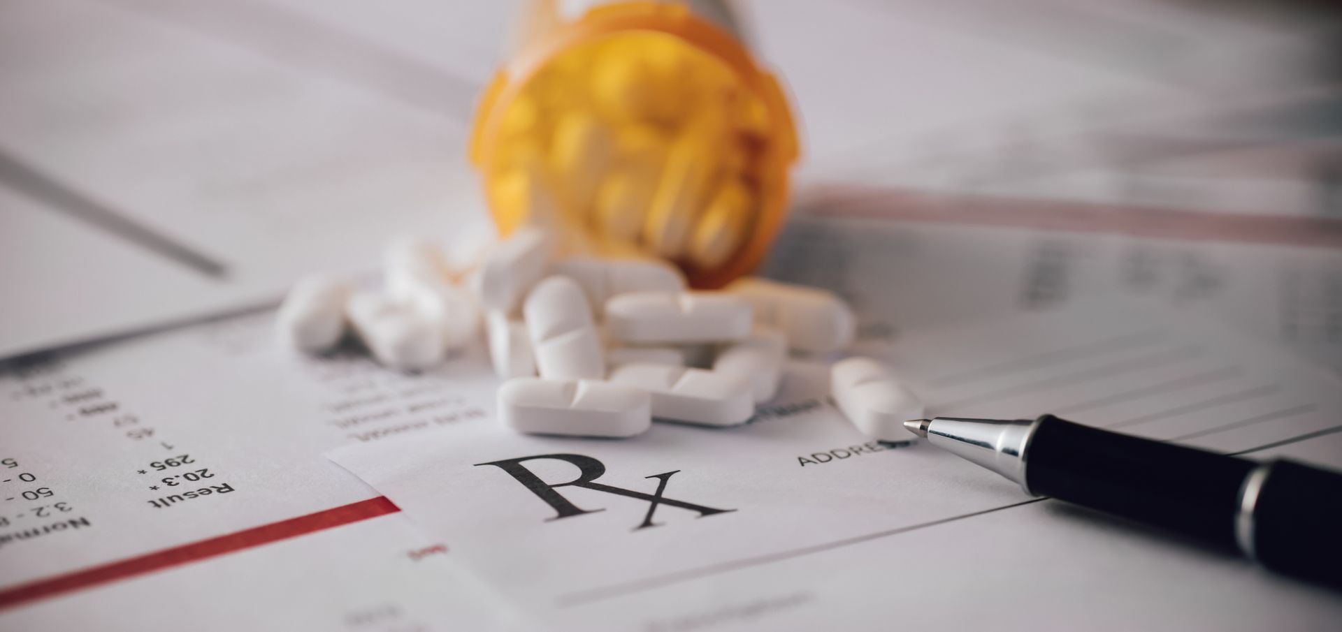 A bottle of pills is spilling out of it on a prescription form with a pen.