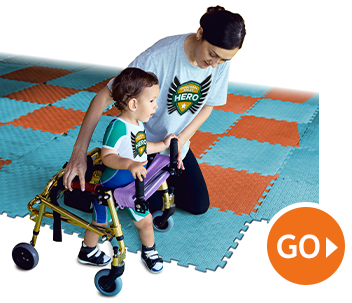 Therapist assisting child in a walker on foam play mats, with a “GO” button icon. Therapist assisting child in a walker on foam play mats, with a “GO” button icon.