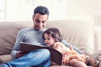 Father and child reading a book together on a couch. The child leans against the father.
