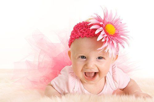 Baby wearing a pink headband with a large flower, lying on a fuzzy surface, smiling.