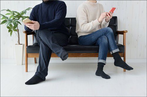 A couple sitting apart on a black couch, looking at their phones. Interior setting with a plant.