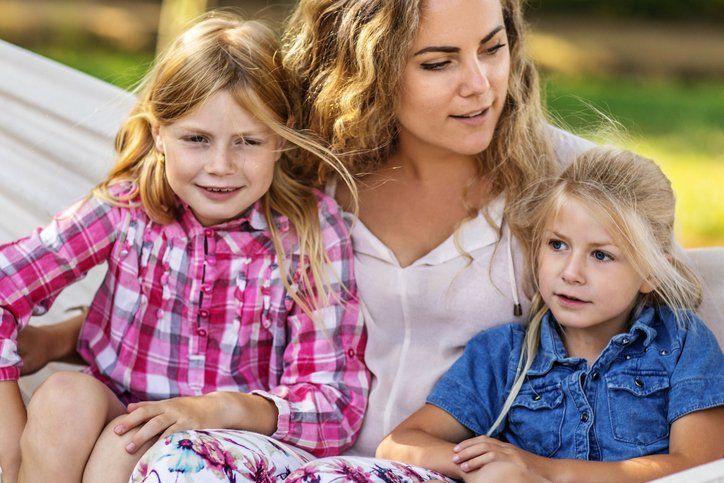 Woman with two young girls seated on a bench outside, reading together.