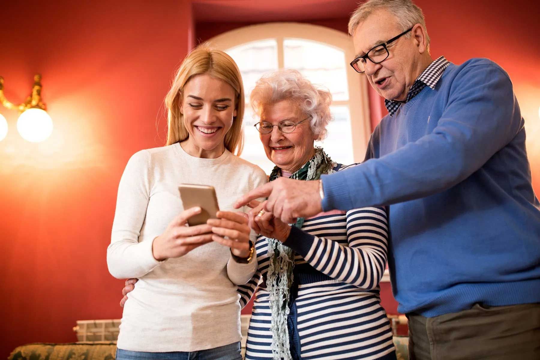 Virtual Visitation — Wilmington, NC — Tise Allan Brandon Woman shows phone to smiling elderly couple, indoors. They point to the screen.